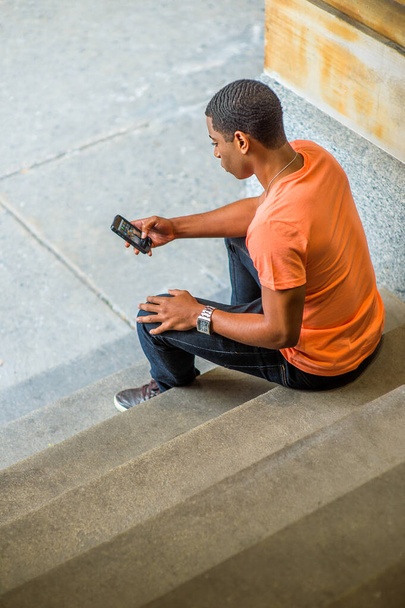 Молодой красивый черный парень сидит на лестнице на углу и проверяет сообщения на мобильном телефоне. - Фото, изображение