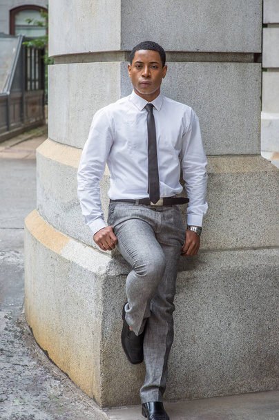 Fehér ingben, fekete nyakkendőben, szürke nadrágban és bőrcipőben, egy fiatal, jóképű fekete egyetemista áll egy kőfal mellett, magabiztosan bámulva téged. - Fotó, kép