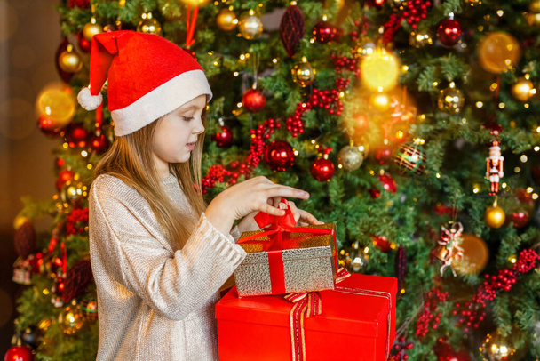 A girl in a Santa hat looks at the gifts at the Christmas tree with a smile - Fotoğraf, Görsel