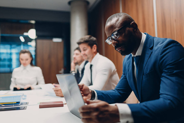 Afričtí američtí černošky muž podnikatel sedí u stolu s notebookem na kancelářském týmu lidé v pozadí. - Fotografie, Obrázek