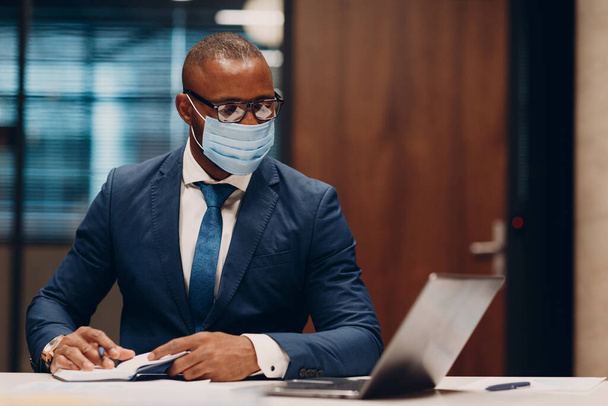 Portrait smiling african american businessman in medical face mask sit at table for meeting in office with notebook with pen and laptop. - Fotó, kép
