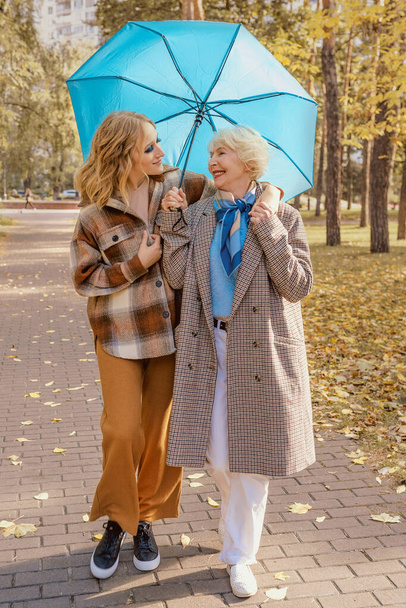 starší žena s mladou dcerou procházející venku pod modrým deštníkem v podzimním parku. Rodina, generace, péče, láska, roční období, koncepce očkování - Fotografie, Obrázek