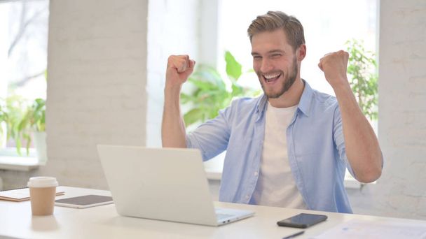 Úspěšný mladý muž slaví na notebooku v moderní kanceláři  - Fotografie, Obrázek