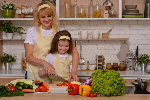 Happy mom teaches daughter to cook (cut vegetables with a knife) at home on the background of the kitchen. Happy pastime, upbringing. Healthy food - Photo, Image
