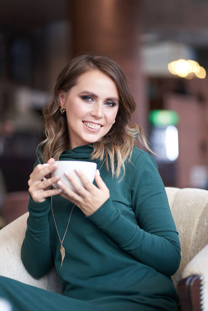 Bir fincan kahve tutan, saç stili olan güzel bir kadın, bir kafede oturuyor. Gülümseyerek doğrudan kameraya bakıyor. - Fotoğraf, Görsel