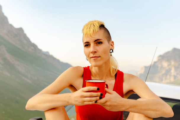 Šťastná mladá dívka kavkazského původu sedí na střeše svého auta, drží červený šálek kávy a dívá se na horolezce, usmívá se a přemýšlí. - Fotografie, Obrázek