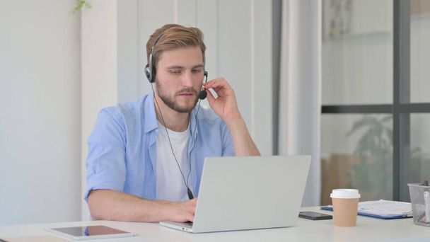 Человек разговаривает на наушниках, работает над ноутбуком в офисе  - Фото, изображение