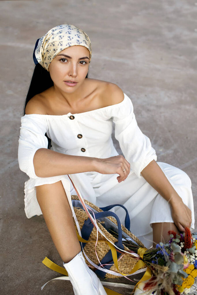 Siedzi stylowa młoda kobieta z szalikiem na głowie i w białej sukience. Piękna dziewczyna z bukietem suszonych dzikich kwiatów i eko-torbą. Pionowe zdjęcie z modnym modelem - Zdjęcie, obraz