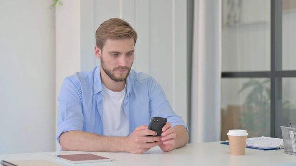 Молодий чоловік використовує смартфон в офісі
  - Фото, зображення