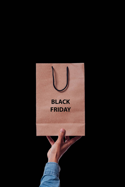 A hand of a man from below holding a shopping paper bag on black background and with a text that says Black Friday - Foto, Imagen