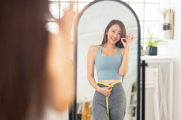 Mladá žena měří velikost pasu před zrcadlem.S gestem OK. Fitness. - Fotografie, Obrázek