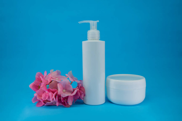 Bílá láhev a sklenice, kosmetické výrobky na modrém pozadí s růžovým květem. Minimální koncept. Showcase pro prezentaci produktů pro péči o pleť - Fotografie, Obrázek