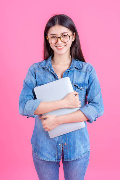 ライフスタイルの美しいアジアのビジネスの若い女性がピンクの背景に隔離されたオンラインホームオフィスのためのラップトップコンピュータを保持-オンラインショッピングとオンラインeラーニングの概念 - 写真・画像