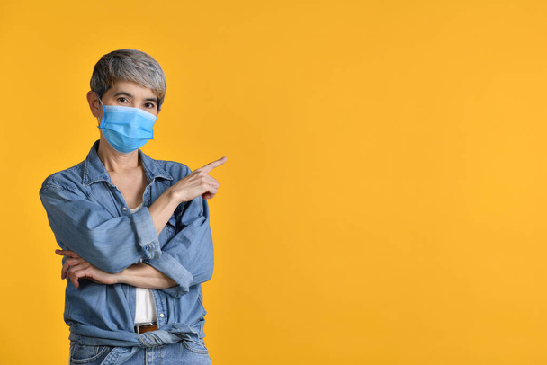 Mulher asiática de meia-idade feliz vestindo máscara cirúrgica para o vírus corona e apontando os dedos para cima no espaço de cópia vazio para texto sobre fundo amarelo - Foto, Imagem