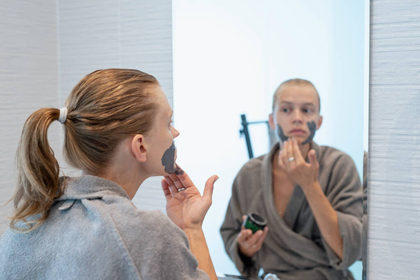 Womens health. Spa and wellness. Woman in gray bathrobes applying face mask in the bathroom looking at the mirror - 写真・画像