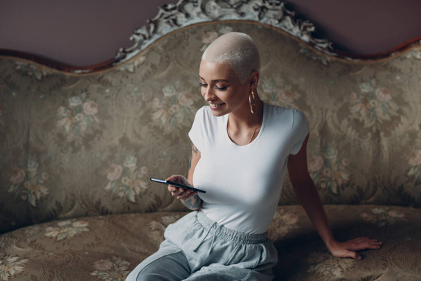Mujer joven millenial con retrato de pelo corto y rubio sentado con teléfono móvil en un sofá vintage. - Foto, Imagen