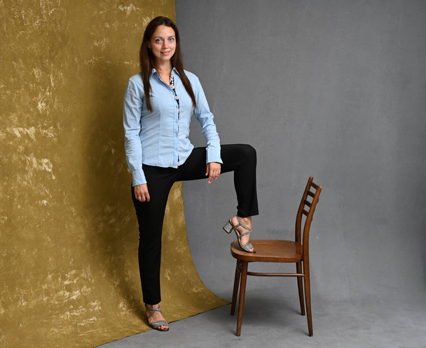 high resolution photo in full growth of a slender woman with a smile in a blue shirt and black trousers standing next to a chair on a gray-beige background - Foto, Bild
