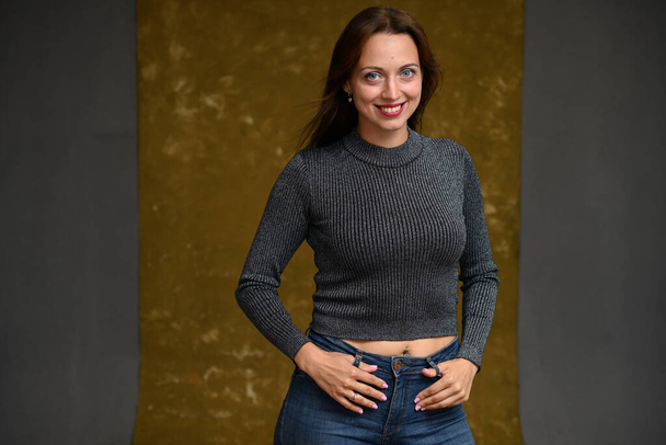 милая улыбающаяся юная стройная женщина с развевающимися волосами в сером свитере позирует в студии на серовато-бежевом фоне - Фото, изображение