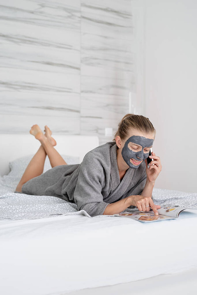 Υγεία των γυναικών. Σπα και ευεξία. Γυναίκα με μάσκα προσώπου χαλαρωτική ξαπλωμένη στο κρεβάτι διαβάζοντας ένα περιοδικό - Φωτογραφία, εικόνα