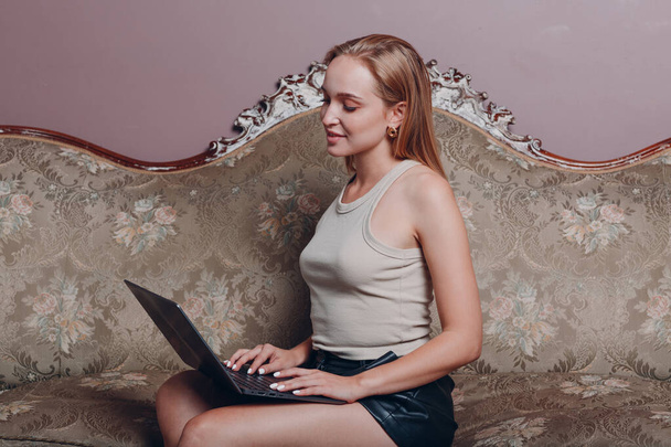 Νεαρή γυναίκα με ξανθά μαλλιά κάθεται και να χρησιμοποιήσετε φορητό υπολογιστή στον καναπέ - Φωτογραφία, εικόνα