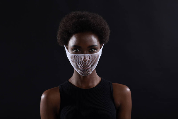 Primo piano ritratto di donna afro-americana modello di moda indossando quarantena viso medico maschera rete grossolana su sfondo nero. Covid-19 concetto di protezione coronavirus. - Foto, immagini