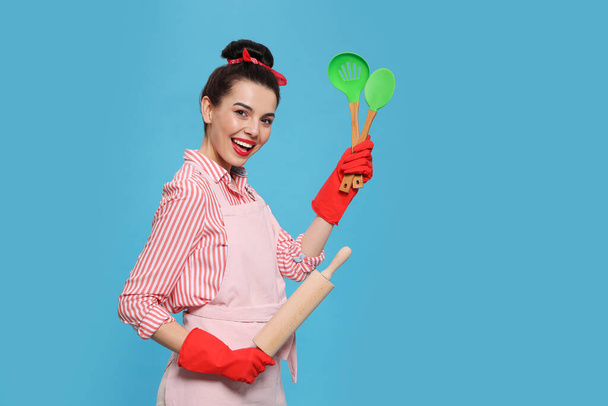 Young housewife with cooking utensils on light blue background - Fotoğraf, Görsel