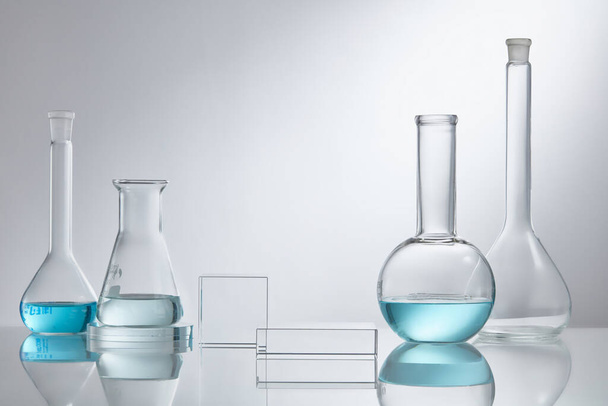 Scena prezentuje kosmetyki na szklanym cokole nowoczesnym w sprzęcie laboratoryjnym. Sprzęt laboratoryjny ze szkła z niebieskimi składnikami wody na białym tle. Koncepcja kosmetyków badawczo-rozwojowych - Zdjęcie, obraz