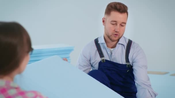 άνδρας με ρούχα εργασίας συζητά με νεαρή γυναίκα styrofoam για μόνωση δαπέδου πριν από την τοποθέτηση laminate στο διαμέρισμα - Πλάνα, βίντεο