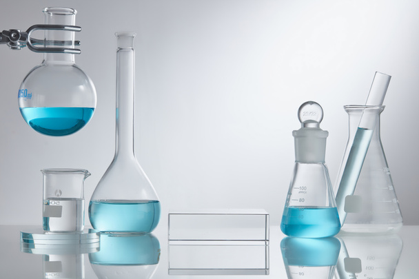 Bühne präsentiert Kosmetik auf Glaspodest modern in der Laborausstattung. Laborglasausrüstung mit blauen Wasserinhaltsstoffen auf weißem Hintergrund. Kosmetikkonzept für Forschung und Entwicklung - Foto, Bild