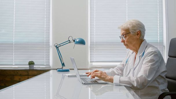 Senior krótkie siwe pani doktor pisze list na współczesnym laptopie siedzi przy stole w przestronnej klinice - Zdjęcie, obraz