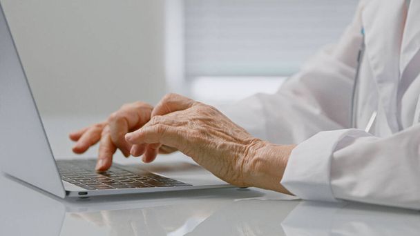 Érett női egészségügyi dolgozó típusok szöveg laptopon ül asztalnál tágas klinika iroda összpontosít ráncos kezek - Fotó, kép