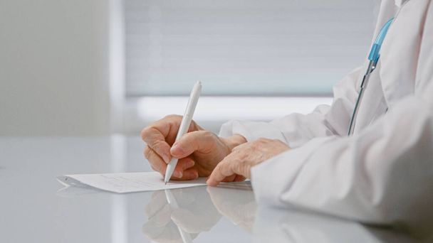 Kıdemli kadın doktor, parlak ışıklandırılmış klinik ofisinde beyaz masada oturan kâğıt üzerinde ilaç reçetesi yazıyor. - Fotoğraf, Görsel