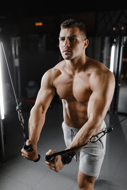 μοντέλο bodybuilder τύπος με γυμνό κορμό, τρένα στο γυμναστήριο. Οι μύες είναι σφιγμένοι. Σκούρο φόντο. - Φωτογραφία, εικόνα