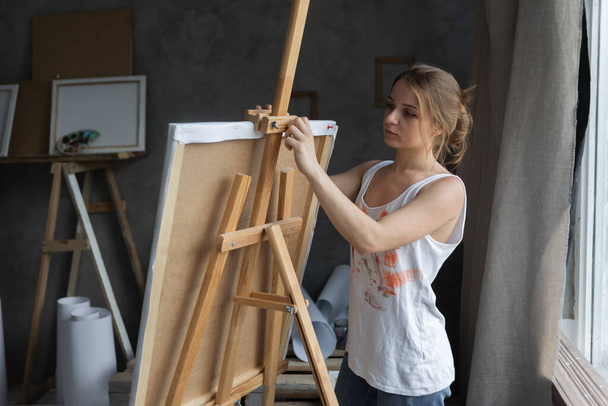 Portrait of a woman artist in an art studio stands near the easel and fixes the canvas. Preparation for drawing. - Fotó, kép