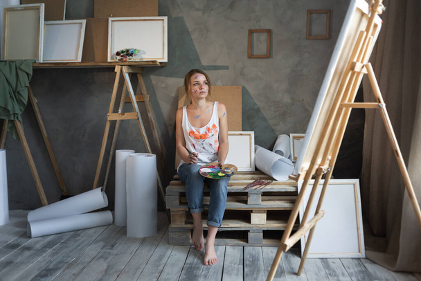 A beautiful Caucasian woman artist sits in a university class near an easel with a canvas holding a palette with mixed paints in her hands and thinks. Copy space. Copy space. - Fotografie, Obrázek