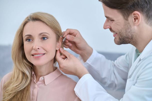 Médico agradável alegre que fixa uma ajuda surda atrás da orelha das mulheres - Foto, Imagem