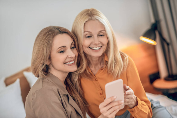 Two smiling blonde women sitting on the bed and making selfie on a smartphone - Valokuva, kuva