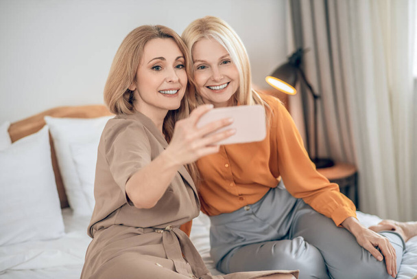 Two smiling blonde women sitting on the bed and making selfie on a smartphone - Фото, изображение