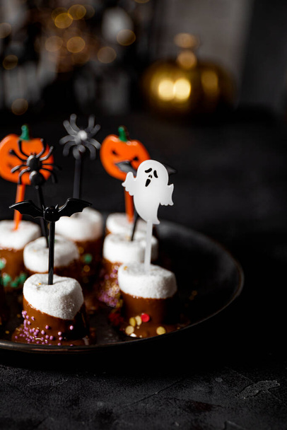  Sweet Halloween treat, chocolate covered marshmallows with sugar and Halloween decorations on a black plate, vertical photo with soft focus - Zdjęcie, obraz