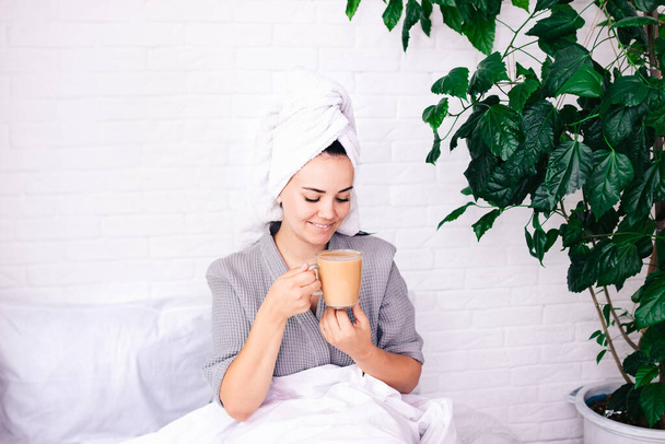 Miellyttävä aamu. Iloinen, viehättävä nuori nainen istuu sängyssä ja juo kahvia. aamunkoiton käsite - Valokuva, kuva