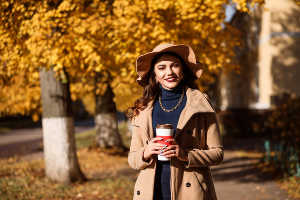 Stylish woman dressed in beige coat, wide brimmed hat, dark blue roll neck sweater and golden chain holds a thermocup with coffee. Outdoors in the city streets in autumn.  - Foto, immagini