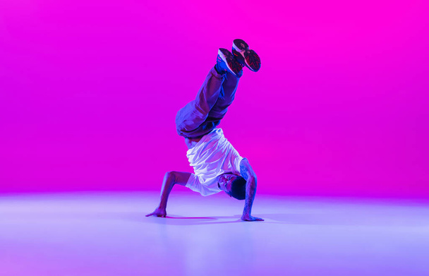 One young stylish man, break dancing dancer training in modern clothes isolated over bright magenta background at dance hall in neon light. - Фото, зображення