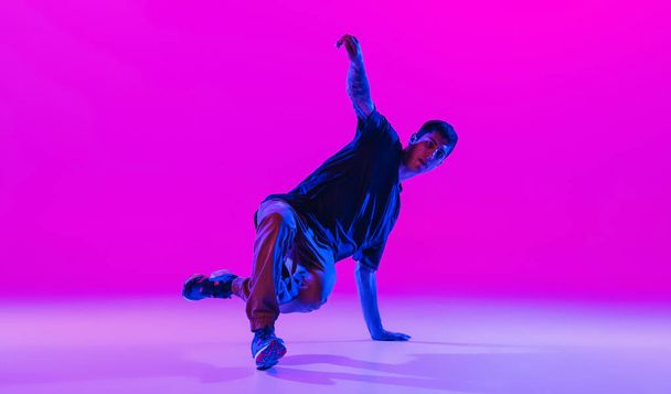 Young stylish man, break dancing dancer practicing solo in modern clothes isolated over bright magenta background at dance hall in neon light. - Photo, image