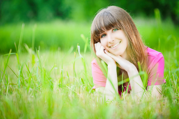 Młoda uśmiechnięta piękna kobieta w czerwonej bluzce leży na trawie na tle zielonego letniego parku. - Zdjęcie, obraz