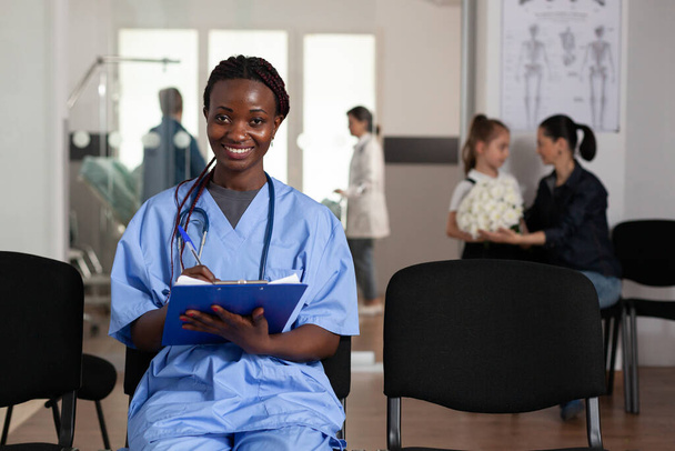 Portrait of african american nurse sitting on chair in hospital waiting area - 写真・画像