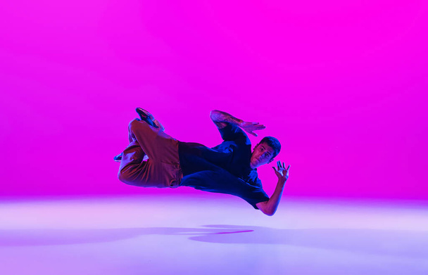 Young stylish man, break dancing dancer practicing solo in modern clothes isolated over bright magenta background at dance hall in neon light. - Фото, зображення
