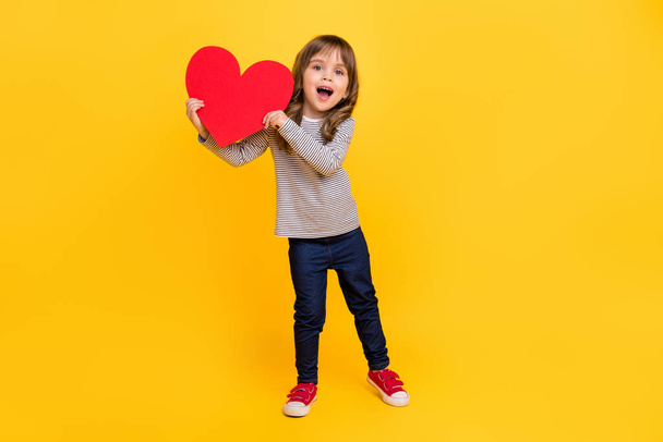 Full body photo of cute little blond hair girl hold heart wear shirt jeans sneakers isolated on yellow backgound - 写真・画像