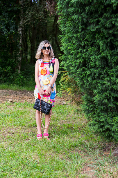 Blondi nainen poseeraa värikäs mekko vihreällä alueella Asturias Espanja.Nainen pukeutuu vaaleanpunainen sandaalit ja musta laukku kädessä.Valokuva on otettu pystysuorassa muodossa aurinkoisena päivänä. - Valokuva, kuva