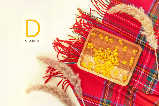 Horizontal banner with vitamin d teme. Autumn composition with wooden plate with yellow vitamin gel capsules, red cashmere palatine and dried flowers on a white background with inscription vitamin d - 写真・画像