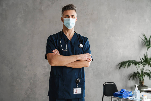 Confident mid aged man doctor wearing medical protective mask looking at camera while standing indoors, arms folded - 写真・画像